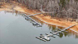 Aerial view of a lakeside area with several interconnected docks extending into the calm water. Five boats are moored at different docks. The shoreline is sandy, bordered by leafless trees. The scene is overcast with a muted color palette.