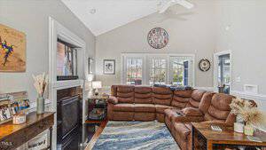 A cozy living room with a brown sectional sofa, a fireplace, and a wall-mounted TV. There are framed photos on a table and a decorative wall clock. Large windows and a door let in natural light, revealing a view of trees outside.