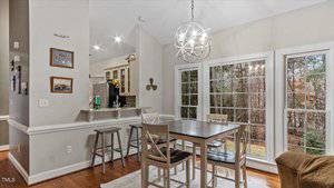 Dining area with a dark wooden table and four chairs. Large windows show a wooded view outside. A modern chandelier hangs from the ceiling. The wall is adorned with framed pictures, and a breakfast bar separates the kitchen and dining space.