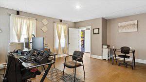 A home office with a desk setup featuring multiple monitors, laptop, and chair. Two windows with beige curtains provide natural light. Additional chairs and a small table are in the room. Walls are beige with a painting and wall decor.