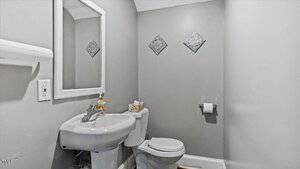 A small bathroom with gray walls features a white pedestal sink, a matching toilet, and a roll of toilet paper on the holder. Above the sink is a rectangular mirror. Decorative square tiles are mounted on the wall next to the toilet.
