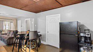 Small kitchen area with a black refrigerator and a countertop holding a coffee maker. A round wooden table with four tall chairs is nearby. The room has a wooden ceiling and a door painted white. The background leads into a living room with a couch.
