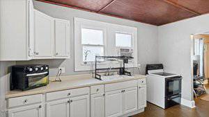 A kitchen with white cabinets and light beige countertops. It features a countertop toaster oven, a drying rack, a window air conditioner, and a stove. The flooring is dark wood, and the ceiling has a wood panel finish.
