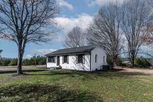 Nestled on 55 Daniel Harris Road in Henderson, this single-story white brick house features a dark roof and black shutters. It sits in an open grassy yard with a small porch, surrounded by leafless trees beneath the partly cloudy sky.