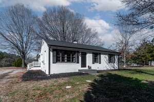 A small white brick house with a black roof and black shutters sits in a yard with sparse grass. A few large leafless trees are in the background. The sky is partly cloudy.