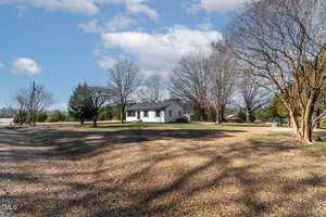 A small white house with a dark roof is surrounded by leafless trees and a grassy yard under a partly cloudy sky. A paved driveway leads towards the house from the left side of the image.