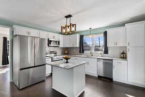 Modern kitchen with white cabinets, granite countertops, and stainless steel appliances, including a refrigerator, oven, and dishwasher. An island in the center holds a fruit bowl. A pendant light hangs above, and curtains frame the window.