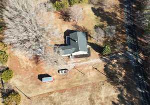 Aerial view of a single-story house surrounded by grass and trees. A driveway leads to the house with a parked truck and a small trailer or shed nearby. A road runs along the right side of the image, bordered by more trees.
