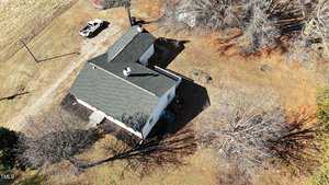 Aerial view of a white house with a dark roof surrounded by bare trees. There is a white vehicle parked nearby on the grass. The ground is covered in a mix of dry grass and fallen leaves. Shadows from the trees stretch across the property.
