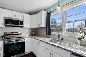 A modern kitchen with white cabinets, a stainless steel oven, and a microwave. There is a double sink under a large window with blue curtains. The backsplash is a textured tile pattern, and a potted plant sits on the counter.