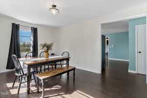 Dining area with a wooden table and benches in a room with white walls and dark wood flooring. Black chairs surround the table, which has a vase with foliage. Large window with black curtains lets in natural light. Adjacent room features a blue wall.