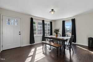A dining room with a rectangular wooden table, six black chairs, and a wooden bench. A potted plant is on the table. Two windows with dark curtains allow natural light. A white door is on the left and hardwood floors cover the room.