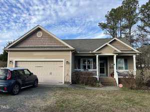 Located at 2616 Pauline Oaks Drive in Franklinton, this single-story house features a beige facade, two-car garage, and welcoming front porch. A black car sits in the driveway, while leafless bushes border the yard against a backdrop of tall pine trees beneath a partly cloudy sky.