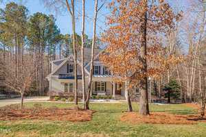 A two-story house with a stone and beige facade is surrounded by trees. There is a manicured lawn and a driveway on the left. A tree with brown leaves is in front, and there are evergreen trees in the background under a clear blue sky.