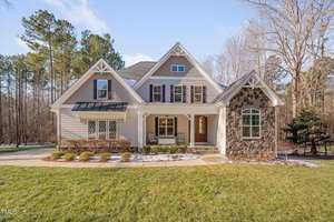 Nestled at 659 Willard Drive, this two-story house in Creedmoor boasts beige siding and stone accents. It features a covered porch with elegant white columns and a dark wooden front door. The peaked roof with dormer windows complements the surrounding trees beneath a clear blue sky.
