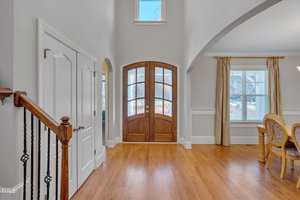 Bright entryway with wooden double doors and arched window above. Adjacent to a dining area with a window and curtains. Hardwood flooring throughout, with a staircase visible to the left and interior doors on the opposite side.