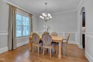 Dining room with light wood floor. Features a wooden table and six upholstered chairs. A chandelier hangs overhead. A window with beige curtains is on the left, and there is an arched doorway leading to another room on the right.