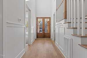 A bright and spacious hallway with wooden flooring leads to double wooden doors with glass panels. Walls are painted white, and a staircase with white railings and wooden steps is visible on the right. There is a room entrance on the left.