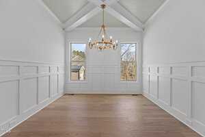 A bright, empty room with white walls and wainscoting features two tall windows and a ceiling with wooden beams. A gold chandelier hangs from the ceiling, and the floor is made of light brown wood. Trees are visible outside the windows.
