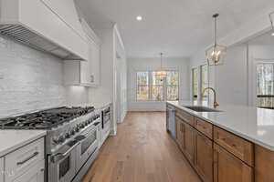 A spacious kitchen featuring a large gas stove and white cabinetry on the left, and a wooden island with a white countertop and sink on the right. Hanging light fixtures illuminate the area, while windows in the background provide natural light.