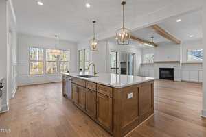 Modern open-concept kitchen and living area featuring a large central island with a white countertop and dark wood cabinetry. The space includes wooden floors, pendant lights, a fireplace, and large windows providing natural light.