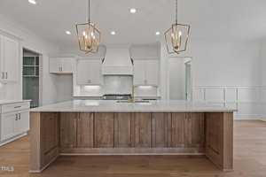 Modern kitchen interior featuring a large center island with wooden paneling and a white countertop. Two geometric pendant lights hang above. The background includes white cabinets, a stove with a hood, and light wood flooring.