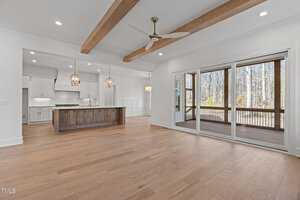 A spacious, modern interior with light wood flooring and a ceiling fan. The open kitchen features an island with pendant lights. Large sliding doors lead to a screened porch with views of a wooded area. White walls and wooden ceiling beams complete the space.