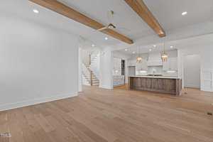 A spacious, empty room with wooden flooring features an open kitchen with a large island, pendant lights, and white cabinets. Exposed wooden beams accent the ceiling. A ceiling fan is installed, and a staircase is visible in the background.