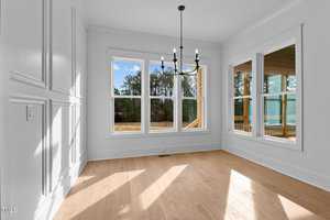 Bright room with light wood flooring and white walls featuring decorative molding. Large windows and a glass door provide views of trees and a patio. A black chandelier hangs from the ceiling, and sunlight streams in, creating shadows on the floor.