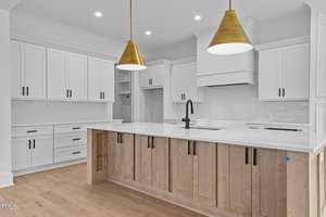 A modern kitchen with white cabinets and a central island featuring light wood panels. Two gold pendant lights hang above the island, and the countertop is white. The faucet is black, and the floor is light wood.