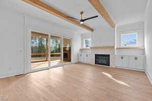 A spacious living room with wooden flooring features a fireplace with a tile surround and built-in cabinets on either side. Large sliding glass doors open to a deck, and a ceiling fan hangs from the ceiling with wooden beams.