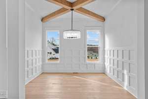 A bright, empty room featuring white walls with a grid pattern, a wooden floor, and a ceiling with exposed wooden beams. Two large windows provide natural light, with a view of houses outside. A modern chandelier hangs from the ceiling.