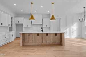 Modern kitchen with white cabinets and a large wooden island featuring a black faucet. Two gold pendant lights hang above the island. The room has wooden flooring, recessed lighting, and a chandelier visible in the adjacent dining area.