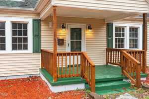 A beige house with green shutters features a small wooden porch with a green painted floor. There are two lantern-style lights flanking the door, and red mulch lines the ground around the porch. The house number 613 is visible.
