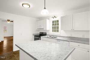 A kitchen with white cabinets, a marble countertop island, and stainless steel appliances. A window above the dual sink allows natural light. In the background, a wooden floor room and a ceiling fan are visible.