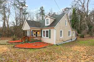 A beige house with a green roof is set in a wooded area. The front has a small porch with a wooden railing. The yard is covered with fallen leaves and features red mulch around the perimeter. Tall trees surround the property.