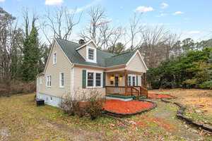 A small house with beige siding and a green roof is situated in a wooded area. It features a front porch with a wooden railing, surrounded by mulch and a stone-bordered pathway. The sky is clear, and trees without leaves are visible in the background.