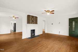 Spacious living room with hardwood flooring, white walls, and a white brick fireplace. Two ceiling fans with wooden blades are visible. A green door is on the right, and a doorway leads to an adjacent room. Another room is visible in the background.
