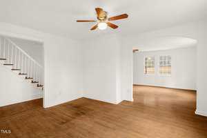 A room with polished wooden floors and white walls. A ceiling fan with a light is in the center. An arched doorway leads to another room with two windows. A staircase with white railings is on the left.