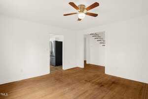 The image shows an empty room with white walls and a wooden floor. A ceiling fan with wooden blades is mounted above. A doorway leads to a kitchen area with a black refrigerator, and another doorway reveals stairs going up.