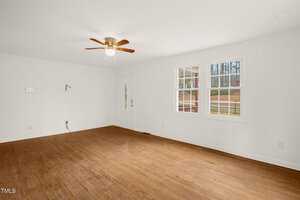 A sparse, empty room with white walls and hardwood flooring. A ceiling fan with a light is centered on the ceiling. Two windows let in natural light, and there is a visible door. Electrical outlets and a wall-mounted fixture are also present.