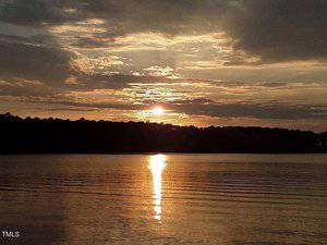 Sunset over a calm lake with a golden sun partially obscured by clouds. The sun's reflection creates a bright line across the water. Dark silhouettes of trees line the horizon under a partly cloudy sky.