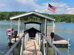 A dock with a covered boat slip extends into a lake at 43 Mt. Pleasant Lane, featuring a boat and two red kayaks. An American flag flies atop the structure. The background includes a treeline and partly cloudy sky as a metal walkway leads to the dock near Manson.
