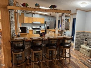 A cozy kitchen and bar area with wooden cabinets and countertops. Four bar stools line the counter. The room features hardwood floors and stone accents. Decorative items, like colorful lizard figurines, are placed above the bar.