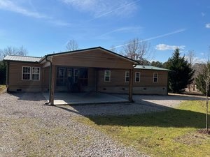 A brown, single-story house with a metal roof is pictured under a clear blue sky. It has a covered porch and driveway with a small grassy area and gravel surrounding it. Leafless trees are seen in the background.
