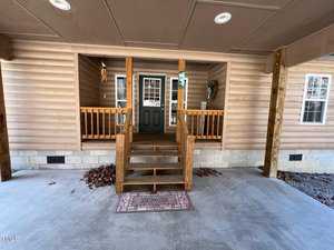 Covered porch entry of a home with wooden siding, featuring a staircase leading to a green front door with glass panels. There are wooden railings on both sides of the stairs, a welcome mat on the concrete floor, and leaves scattered near the sides.