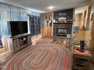 A living room features a large TV on a wooden stand, with corrugated metal walls and patterned curtains. There's a stone fireplace with decorative items above, wooden cabinets, and an oval braided rug on the wood floor. A lamp sits on a side table.
