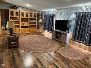 Living room with wooden flooring and walls. A large braided oval rug and a smaller matching rug on the floor. Entertainment center with a TV, bookshelves with decorative items, side table with a lamp, and star-patterned curtains on the windows.