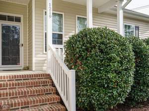 A porch with a set of red brick steps and white railing leading to a dark front door. The house exterior is light-colored siding. A large green bush with small red berries is on the right side of the steps. House number 1219 is visible.