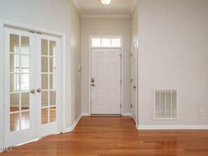 A residential entryway featuring a white front door with a window above it. The floor is polished wood. To the left, there's a set of double glass doors. Walls are painted light gray, and a wall vent is on the right. Ceiling light is centered.